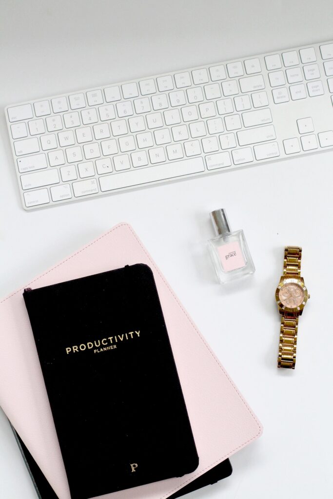 A keyboard with a perfume bottle, watch, and planner sitting on a table.