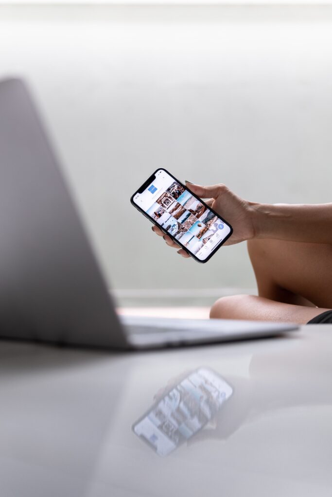 A woman holding a phone looking at a social media platform with a computer next to her. 