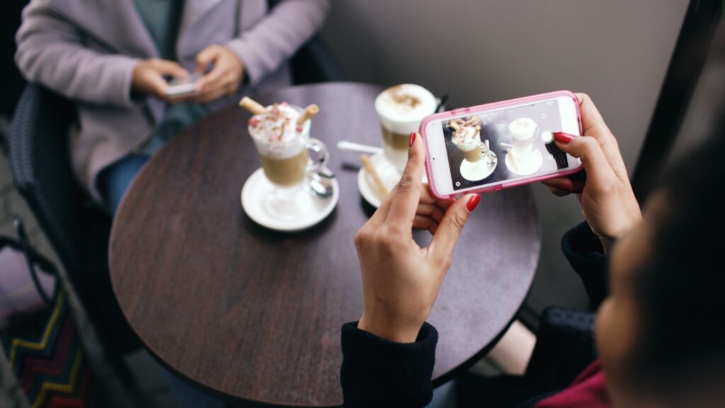 Person having coffee with a friend and taking a picture of the coffee with their phone to post on social media. 